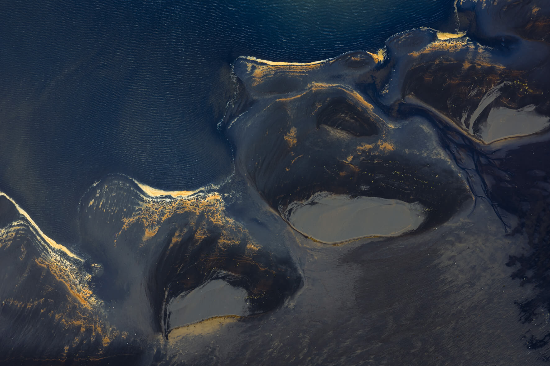 Aerial view of Icelandic lagoon where golden foam traces curve across deep blue tidal water